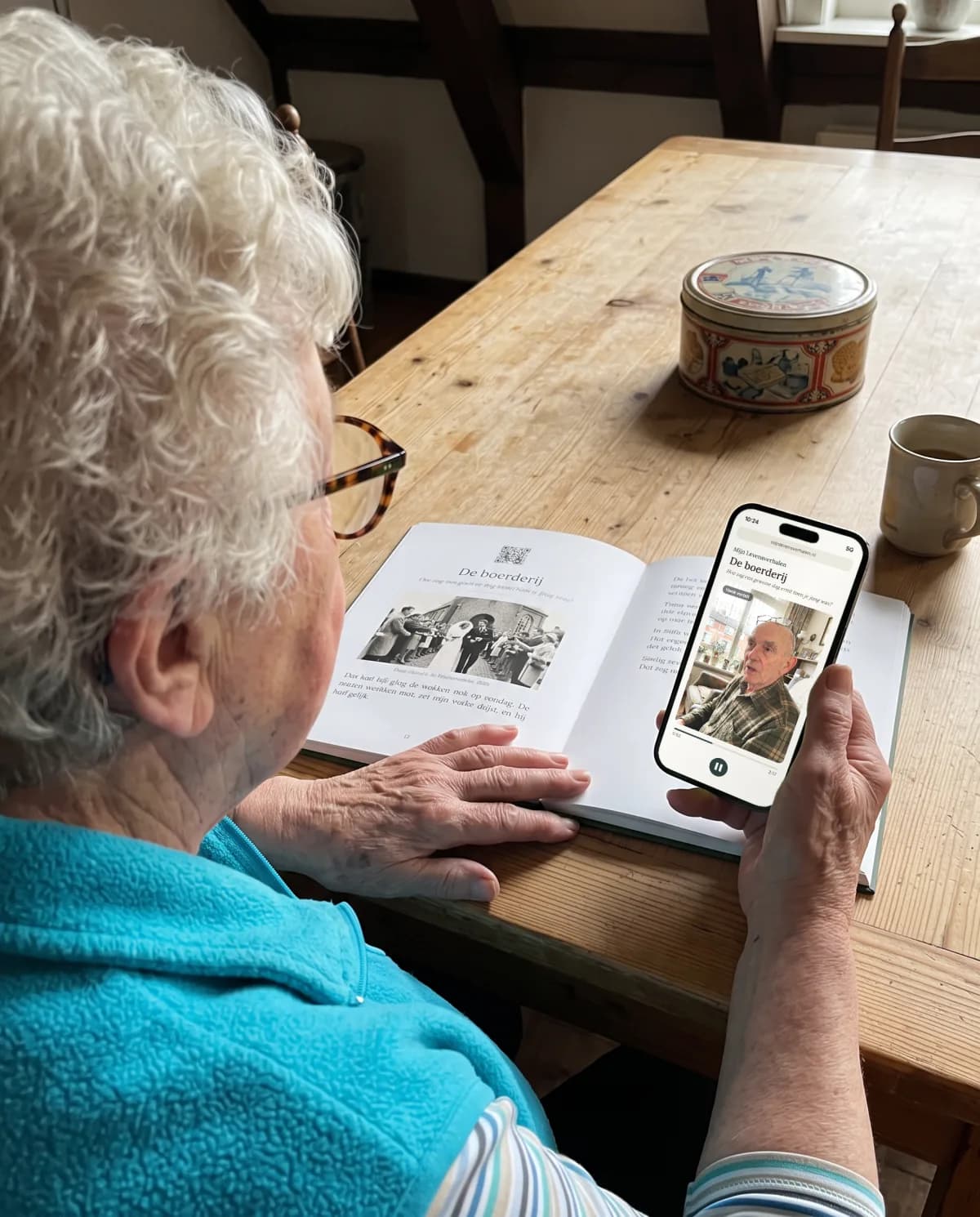 Eine Enkelin hört am Küchentisch auf ihrem Handy Opas Geschichte, sein Buch liegt neben ihr.
